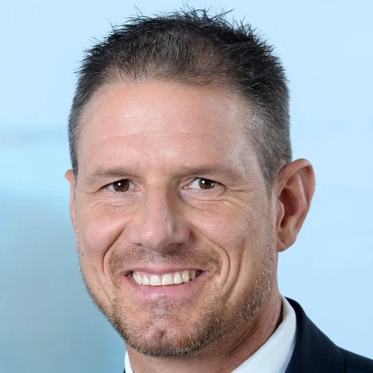 Ein Mann mit kurzen braunen Haaren und einem gestutzten Bart lächelt. Er trägt eine dunkle Anzugsjacke, ein weißes Hemd und eine Krawatte vor einem hellblauen Hintergrund.