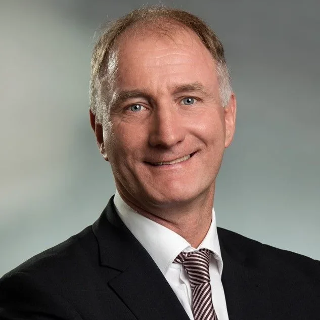 Ein Mann mittleren Alters mit kurzen hellbraunen Haaren trägt einen schwarzen Anzug, ein weißes Hemd und eine gestreifte Krawatte und lächelt in die Kamera vor einem verschwommenen grauen Hintergrund.