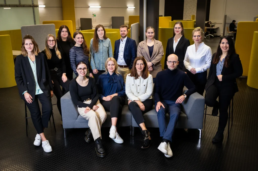 Eine Gruppe von vierzehn professionell gekleideten Personen posiert für ein Foto in einem modernen Büroraum mit gelben Trennwänden und grauen Möbeln. Einige sitzen auf einem Sofa, andere stehen hinter ihnen, alle schauen in die Kamera und lächeln.