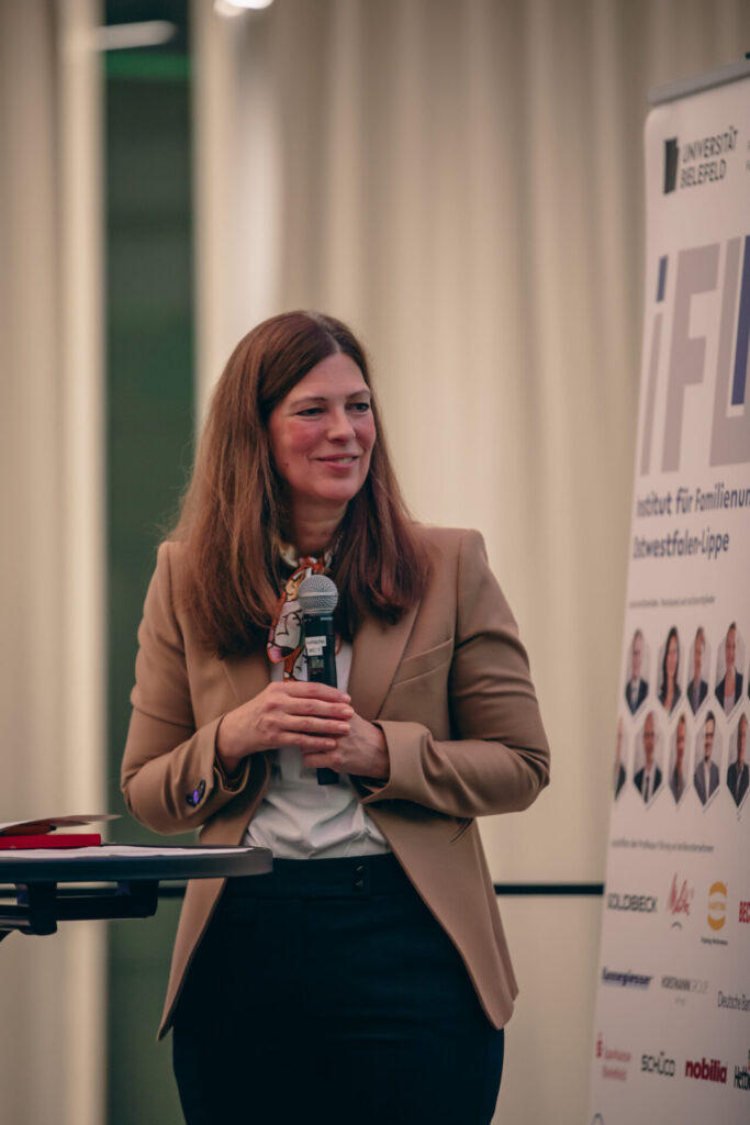 Eine Frau in einem hellbraunen Blazer hält ein Mikrofon und lächelt, während sie bei einer Indoor-Veranstaltung neben einem vertikalen Banner steht. Das Banner zeigt das Logo der Universität Bielefeld und mehrere Firmenlogos.