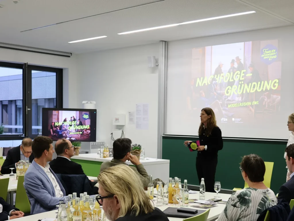 Eine Frau steht vor einem sitzenden Publikum in einem modernen Konferenzraum. Auf einem großen Bildschirm wird eine Präsentation mit dem Titel NACHFOLGE - GRÜNDUNG gezeigt. Auf den Tischen sind Flaschen und Gläser angeordnet.