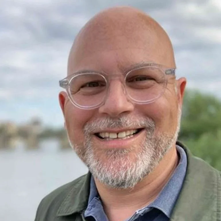 Ein kahlköpfiger Mann mit kurzem grauem Bart und einer Brille mit klarem Gestell lächelt im Freien, mit einem bewölkten Himmel und Grünpflanzen im Hintergrund.