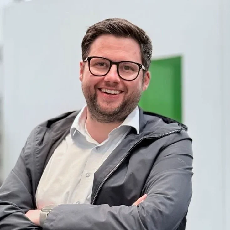 Ein Mann mit kurzen dunklen Haaren und Brille lächelt, während er mit verschränkten Armen steht. Er trägt ein weißes Hemd und eine graue Jacke vor einem hellen Hintergrund mit einer grünen rechteckigen Form.