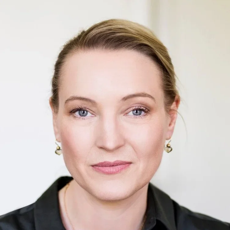 Eine Frau mit heller Haut, blauen Augen und zurückgestecktem blondem Haar, die Perlenohrringe und ein schwarzes Hemd mit Kragen trägt, lächelt sanft vor einem hellen Hintergrund.