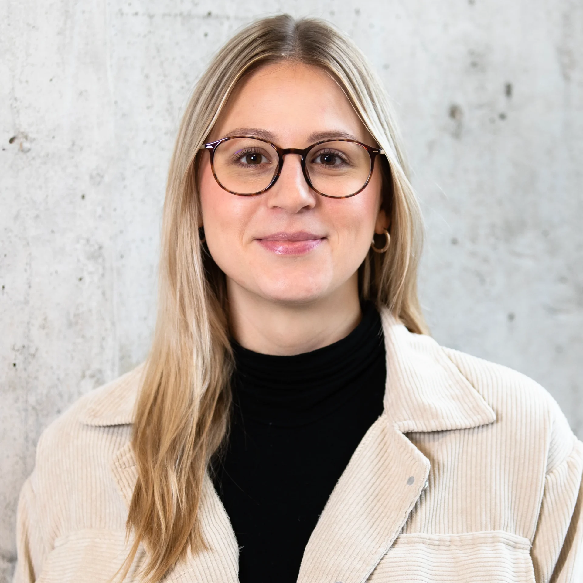 Eine junge Frau mit langen blonden Haaren, die eine runde Brille, einen schwarzen Rollkragenpullover und eine beigefarbene Kordjacke trägt, steht vor einer hellen Betonwand und lächelt sanft in die Kamera.