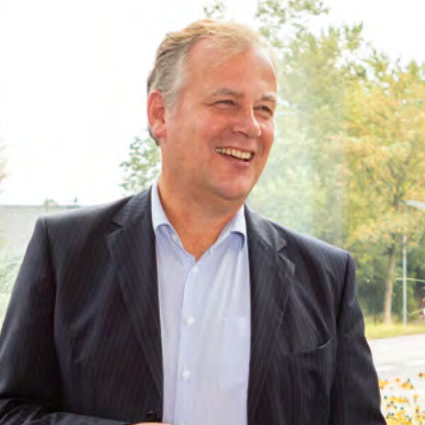 Ein lächelnder Mann in einem dunklen Anzug und einem hellblauen Hemd steht im Freien mit Bäumen und Grünpflanzen im Hintergrund.