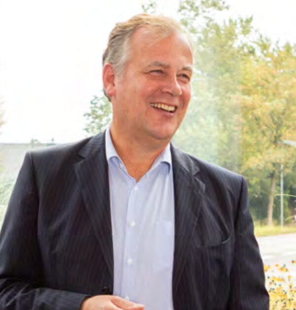 Ein lächelnder Mann in einem dunklen Anzug und einem hellblauen Hemd steht im Freien mit Bäumen und Grünpflanzen im Hintergrund.