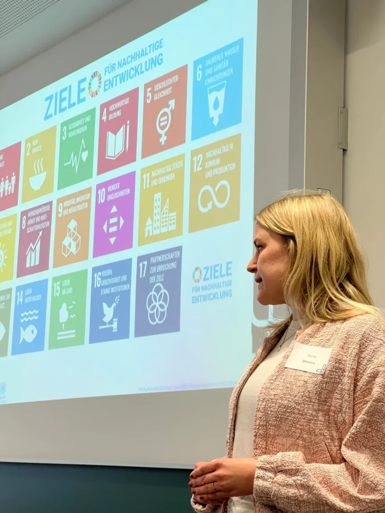 Eine Frau hält einen Vortrag über die Ziele der Vereinten Nationen für nachhaltige Entwicklung, während hinter ihr eine bunte Folie mit den 17 Zielen und ihren Symbolen an die Wand projiziert wird.