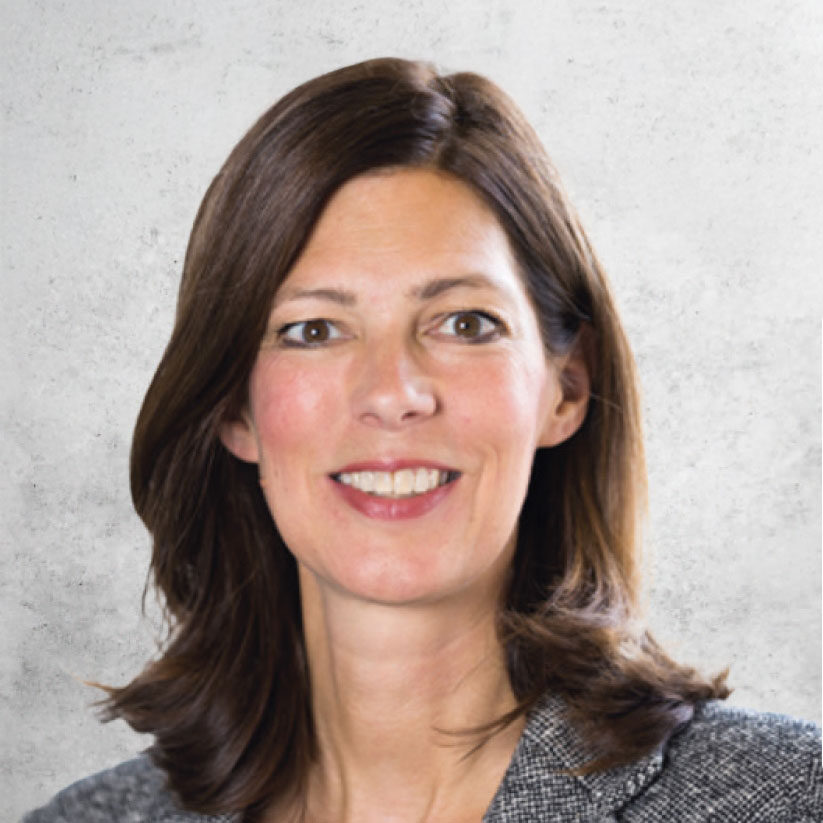 Eine Frau mit schulterlangem braunem Haar, die einen grauen Blazer trägt und lächelt, posiert vor einem hellgrauen, strukturierten Hintergrund.