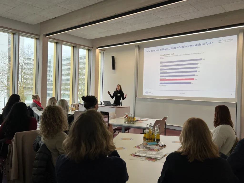 Eine Frau hält vor einer Gruppe von Personen in einem hellen Konferenzraum eine Präsentation. Eine Folie mit einem Balkendiagramm und deutschem Text wird an die Wand projiziert. Die Anwesenden sitzen an Tischen mit Getränken und Papieren. Große Fenster lassen Tageslicht herein.