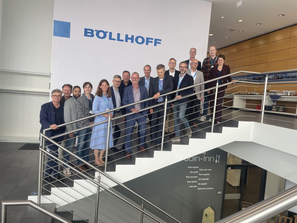 Eine Gruppe von Menschen in Geschäftskleidung steht lächelnd auf einer modernen Treppe in einem Gebäude mit einem großen Böllhoff-Logo an der Wand hinter ihnen. Die Umgebung ist hell und professionell.