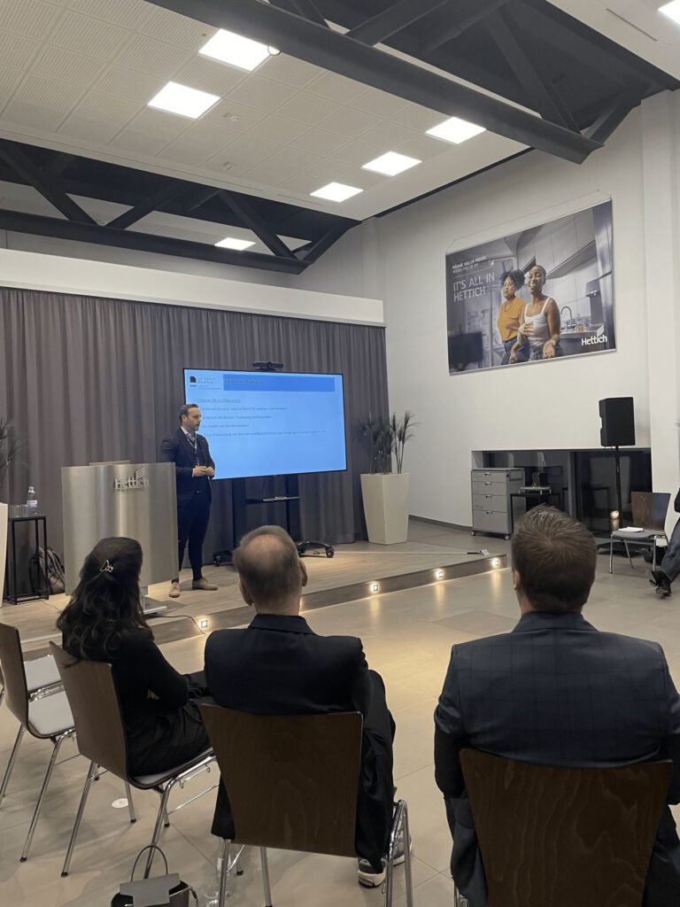 Ein Mann im Anzug hält einen Vortrag vor einem Publikum in einem modernen Konferenzraum. Er steht in der Nähe eines großen Bildschirms, auf dem Dias gezeigt werden, und im Hintergrund sind ein Plakat und industrielle Dekoration zu sehen.