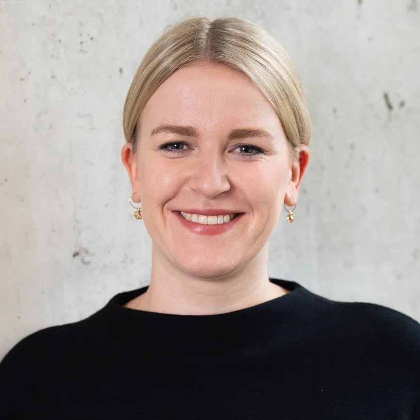 Eine Frau mit blondem, in der Mitte gescheiteltem Haar, die kleine Ohrringe und ein schwarzes Oberteil trägt, lächelt vor einer hellen, strukturierten Wand.