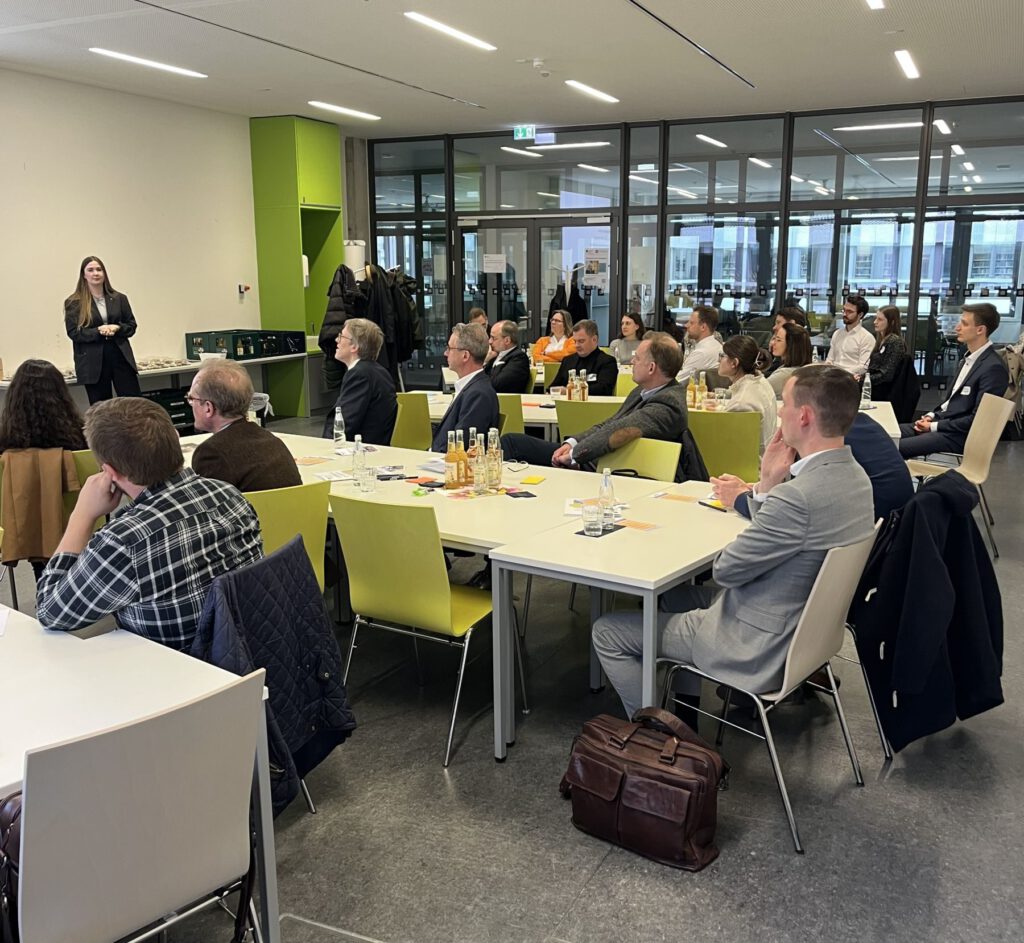 Eine Gruppe von Personen sitzt an Tischen in einem modernen Konferenzraum und blickt auf eine Frau, die an der Stirnseite steht und spricht. Die Zuhörer sehen aufmerksam aus, und auf den Tischen stehen Flaschen, Gläser und Notizblöcke.