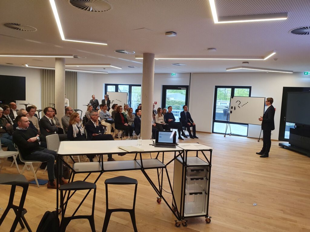 Ein Mann im Anzug hält einen Vortrag vor einem Publikum, das in einem modernen Konferenzraum mit großen Fenstern, Whiteboards und einem Bildschirm sitzt. Einige Leute hören aufmerksam zu, während andere sich Notizen machen.