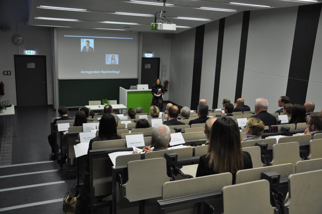 Eine Frau steht an der Stirnseite eines modernen Hörsaals und hält einen Vortrag vor einem Publikum, das in Reihen sitzt. Ein Projektor zeigt Dias mit Text und Fotos auf einer Leinwand über einer Kreidetafel.