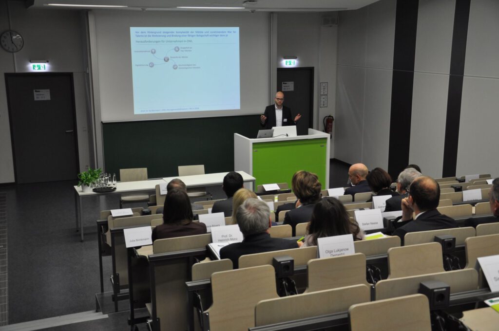 Ein Mann im Anzug hält einen Vortrag vor einem sitzenden Publikum in einem Hörsaal. Er steht an einem Podium mit einer grünen Frontplatte, während hinter ihm ein Dia an die Wand projiziert wird.