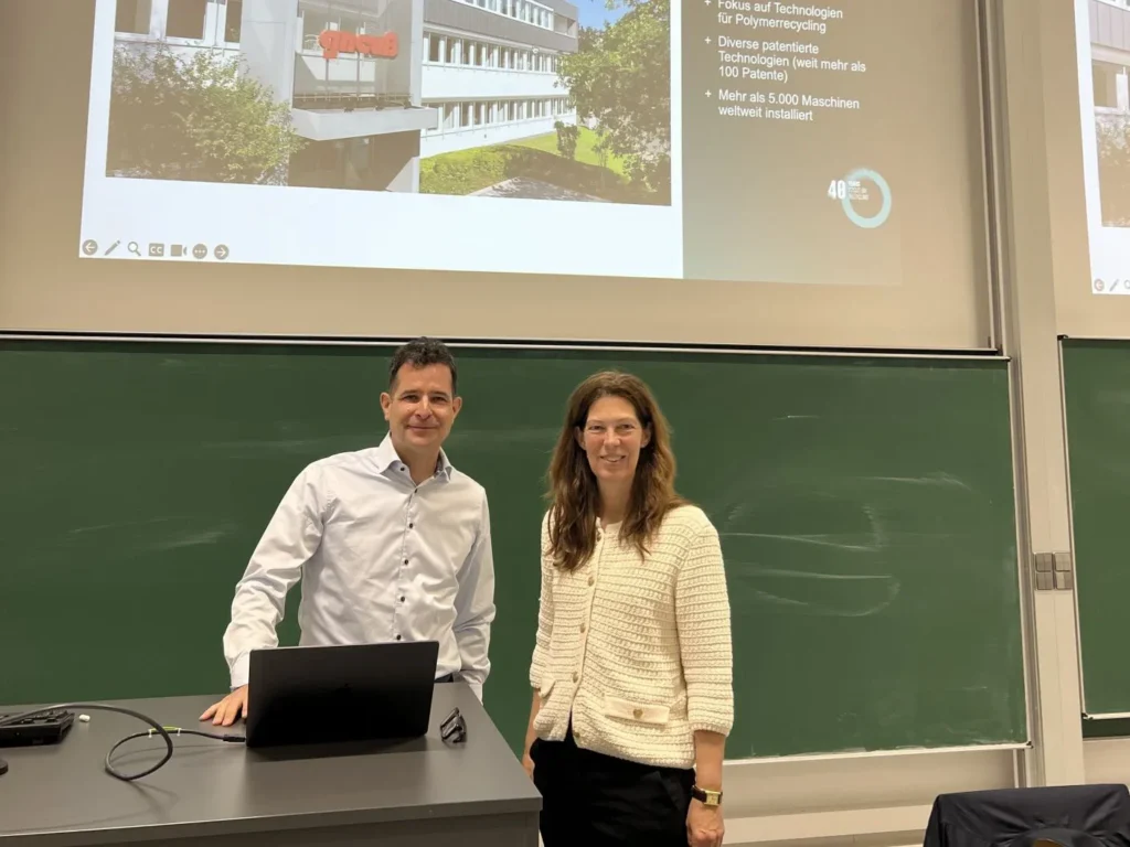 Zwei Personen, ein Mann in einem weißen Hemd und eine Frau in einem cremefarbenen Pullover, stehen lächelnd vor einem Podium in einem Klassenzimmer mit grünen Kreidetafeln. Auf der Leinwand hinter ihnen ist eine Präsentationsfolie zu sehen.