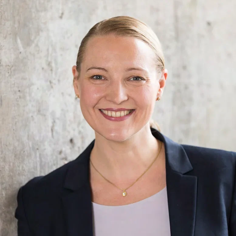 Eine Frau mit zurückgebundenem blondem Haar lächelt in die Kamera. Sie trägt einen dunklen Blazer über einem hellen Oberteil und eine goldene Halskette. Der Hintergrund ist eine strukturierte, helle Wand.