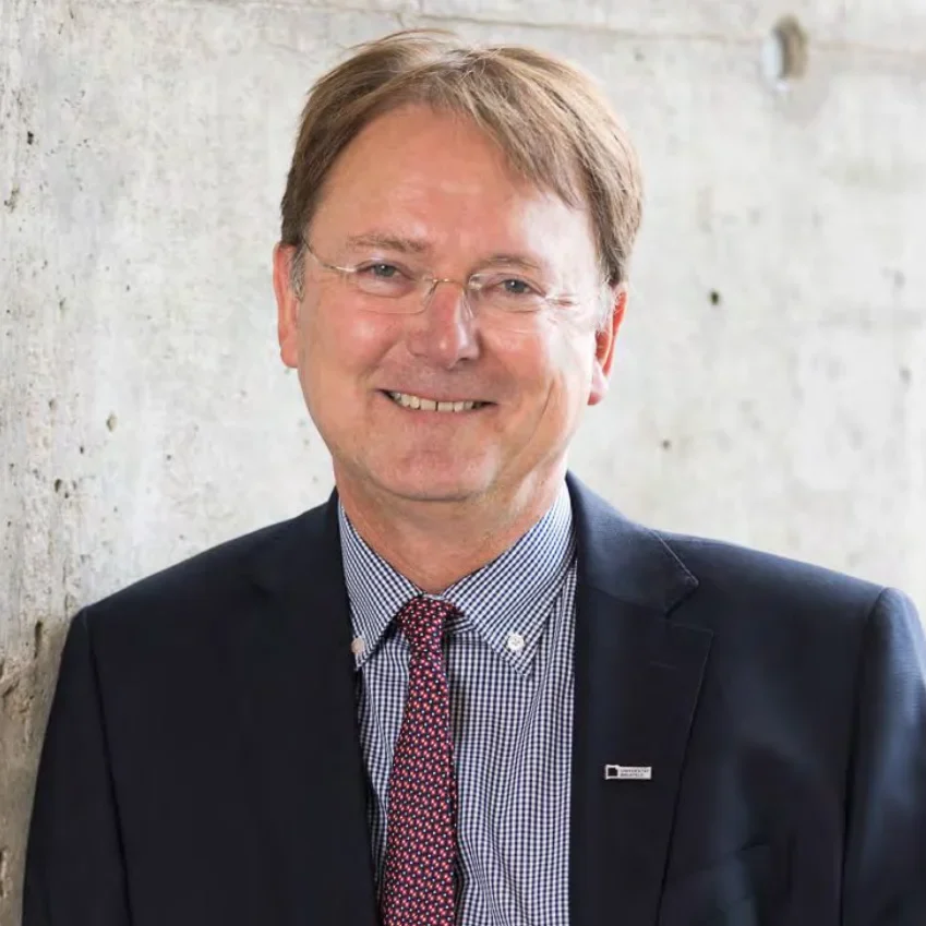 Ein Mann mittleren Alters mit Brille, kurzen braunen Haaren und einem freundlichen Lächeln, der einen dunklen Anzug, ein kariertes Hemd und eine rot gemusterte Krawatte trägt, steht vor einer Betonwand.