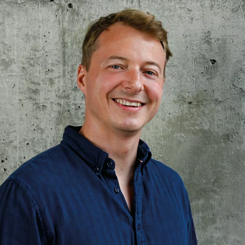 Ein lächelnder Mann mit kurzen hellbraunen Haaren und einem dunkelblauen Button-up-Hemd steht vor einer strukturierten Betonwand.