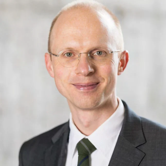 Ein lächelnder Mann mit Brille, heller Haut und schütterem Haar, der einen dunklen Anzug, ein weißes Hemd und eine gemusterte Krawatte trägt, steht vor einem verschwommenen hellen Hintergrund.