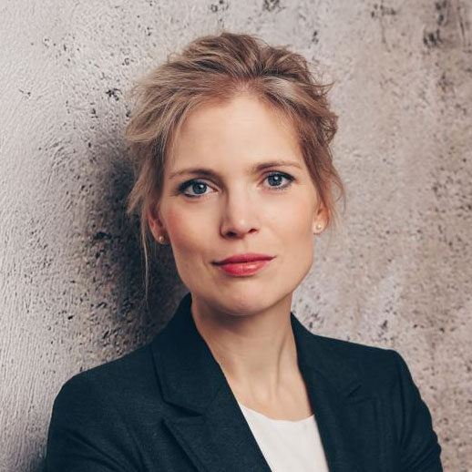 Eine Frau mit blondem Haar, die einen dunklen Blazer und eine weiße Bluse trägt, steht vor einer strukturierten, hellen Wand und blickt mit neutralem Blick direkt in die Kamera.