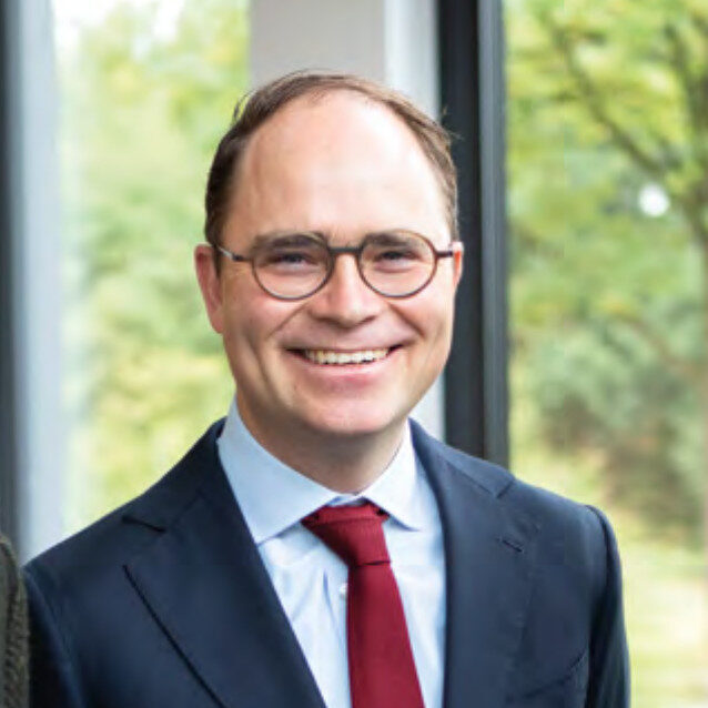 Ein Mann mit Brille, dunklem Anzug, weißem Hemd und roter Krawatte lächelt in einem Haus mit großen Fenstern und grünem Laub im Hintergrund.