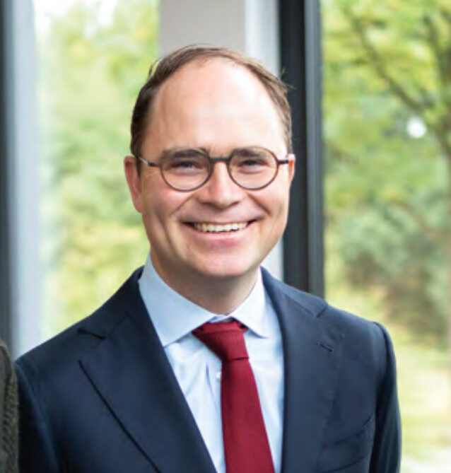 Ein Mann mit Brille, dunklem Anzug, weißem Hemd und roter Krawatte lächelt in einem Haus mit großen Fenstern und grünem Laub im Hintergrund.